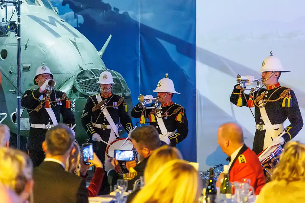 Royal Marines drummers in full ceremonial dress performing with bugles and drums at the Employer Recognition Scheme Gold Awards, with guests watching from seated tables