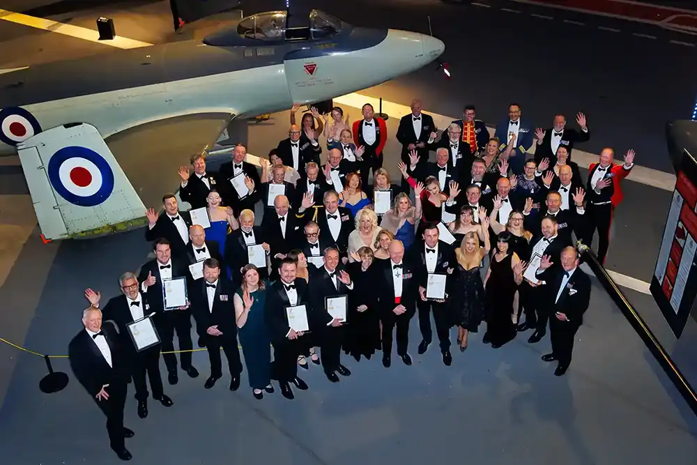 Large group of Gold Employer Recognition Scheme award winners in formal evening wear, standing together beneath an aircraft and holding their certificates while waving at the camera